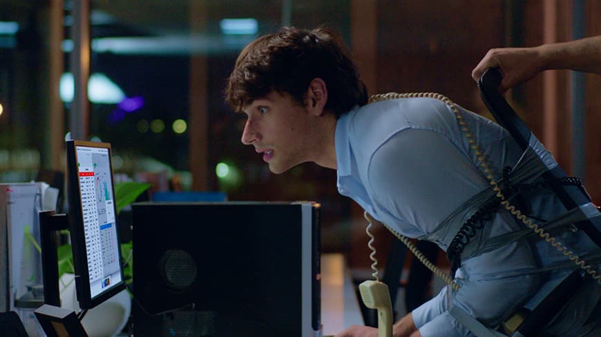 A young office worker is tied to his office chair, he leans forward towards his computer.