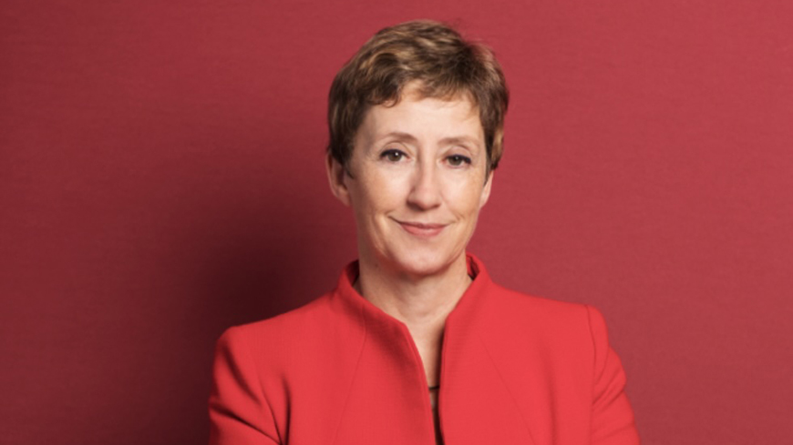 A person with short hair stands confidently with crossed arms, wearing a red blazer over a black top and a checkered skirt, against a plain red background.