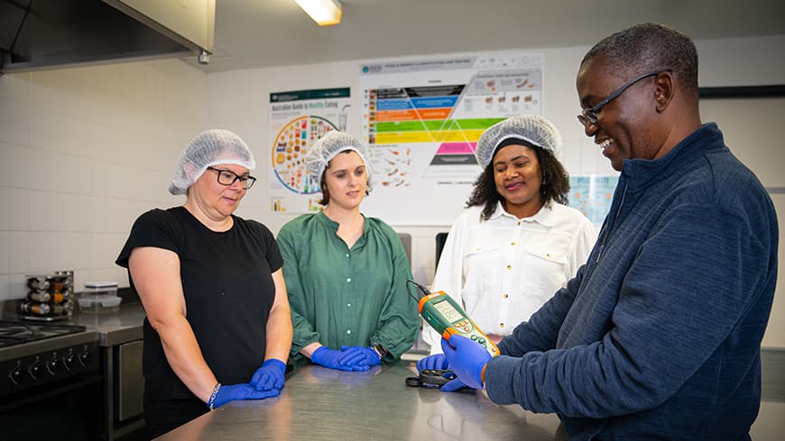 Four people standing around a steel topped bench in an industrial kitchen, three women in white hair nets and man in blue shirt and glasses in profile holding a measurement monitor