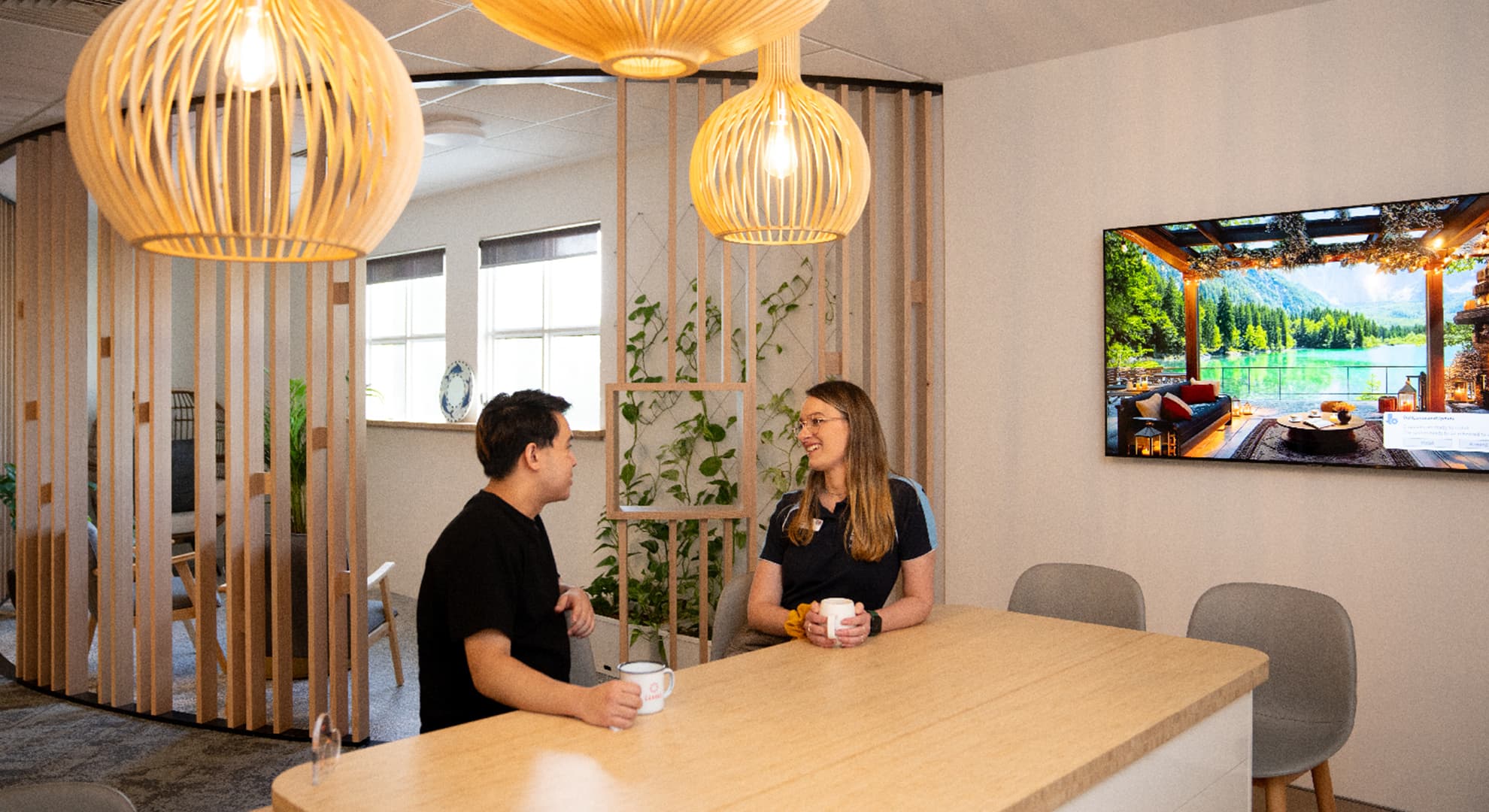 Two people sitting talking at light wooden table with cups of tea, with screen on living room scene to the right and light wood panelled space in background