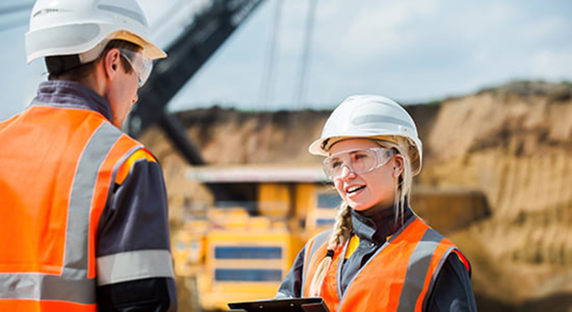 Two highvis mine workers in hard hats talking to each other, man on left with back to camera, young woman with blonde braid over her shoulder and safety glasses with mining vehicle and mine site blurry in the background