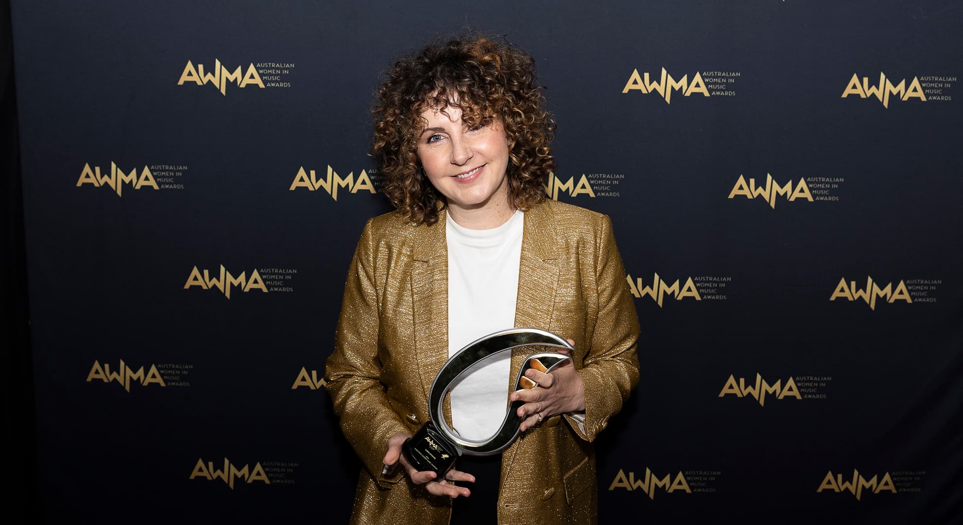 Smiling curly brown haired woman in gold jacket over white t-shirt holding silver award in front of black media wall with AWMA gold logos on it