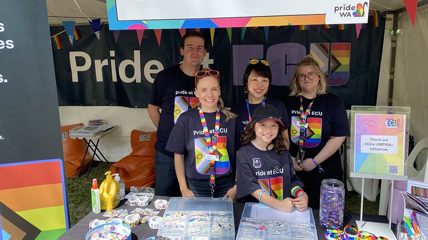 Four people and a child in ECU Pride t-shirts standing in a stall behind desk with Pride merchandise.