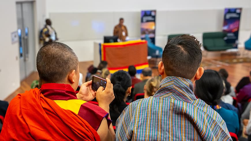 Photograph from behind two heads looking down in a lecture theatre - two people one in traditional Bhutanese monk robe and other in traditional Bhutanese dress