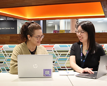 A student is engaged with a Librarian, both meet with a laptop in a spacious Library setting.