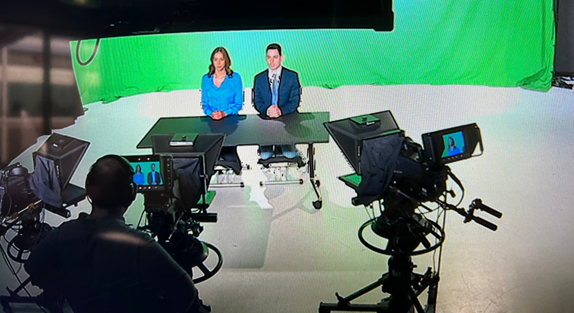 Two ECU Journalism and Broadcast Media students producing a TV program, they are in the middle of filming with two cameras and a green screen behind them.