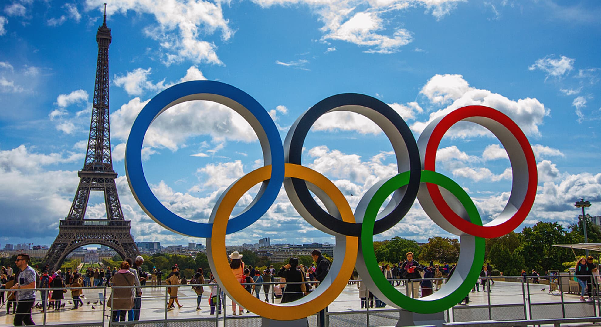 giant coloured olympic rings in front of paved plaza of tourists with Eiffel Tower in background in Paris