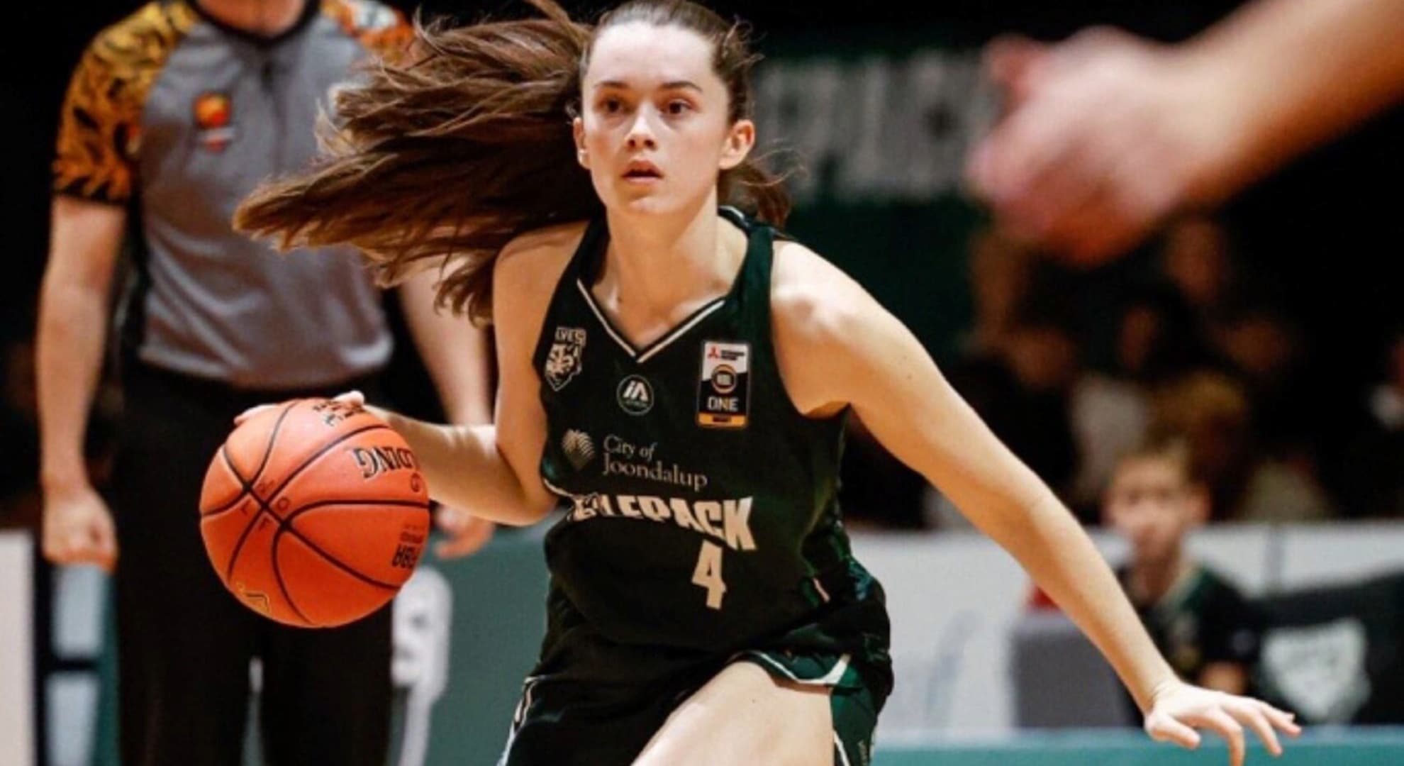 action photograph of young woman playing basketball, black singlet and shorts uniform long brown hair in pony tail caught in action hair flying above her head, umpire standing behind he