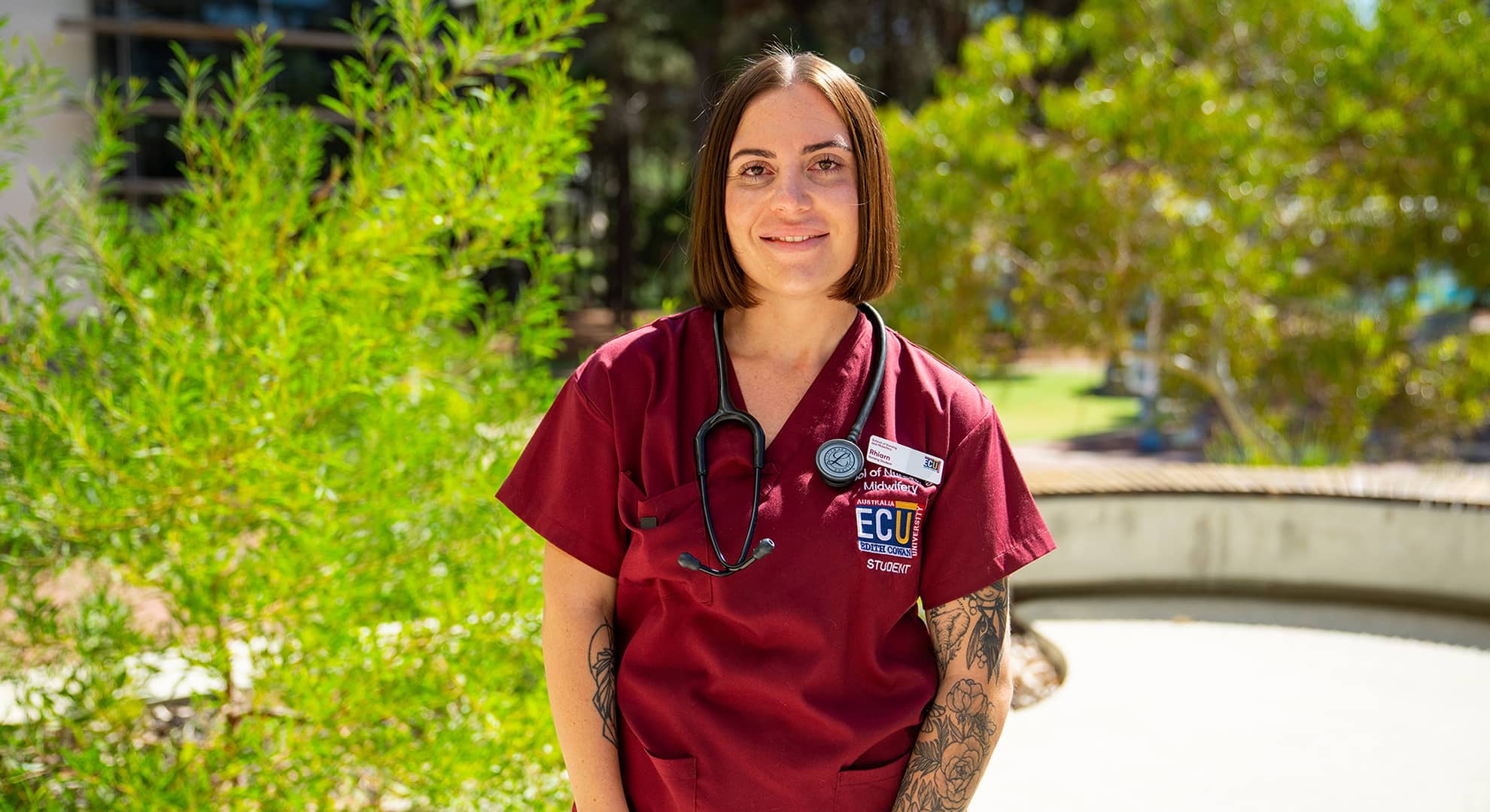 Young woman in maroon scrubs with ECU logo and a stethoscope around her neck and floral tatooes on left arm