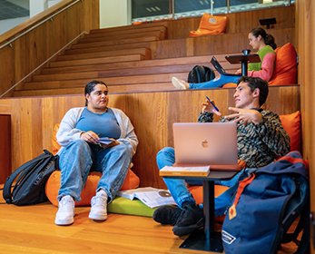 Group of students studying in the library while sitting on library stares area  with laptops and books.