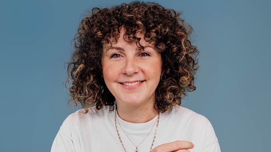 Head and shoulders portrait of smiling curly brown haired woman in white t-shirt in front of grey / blue background