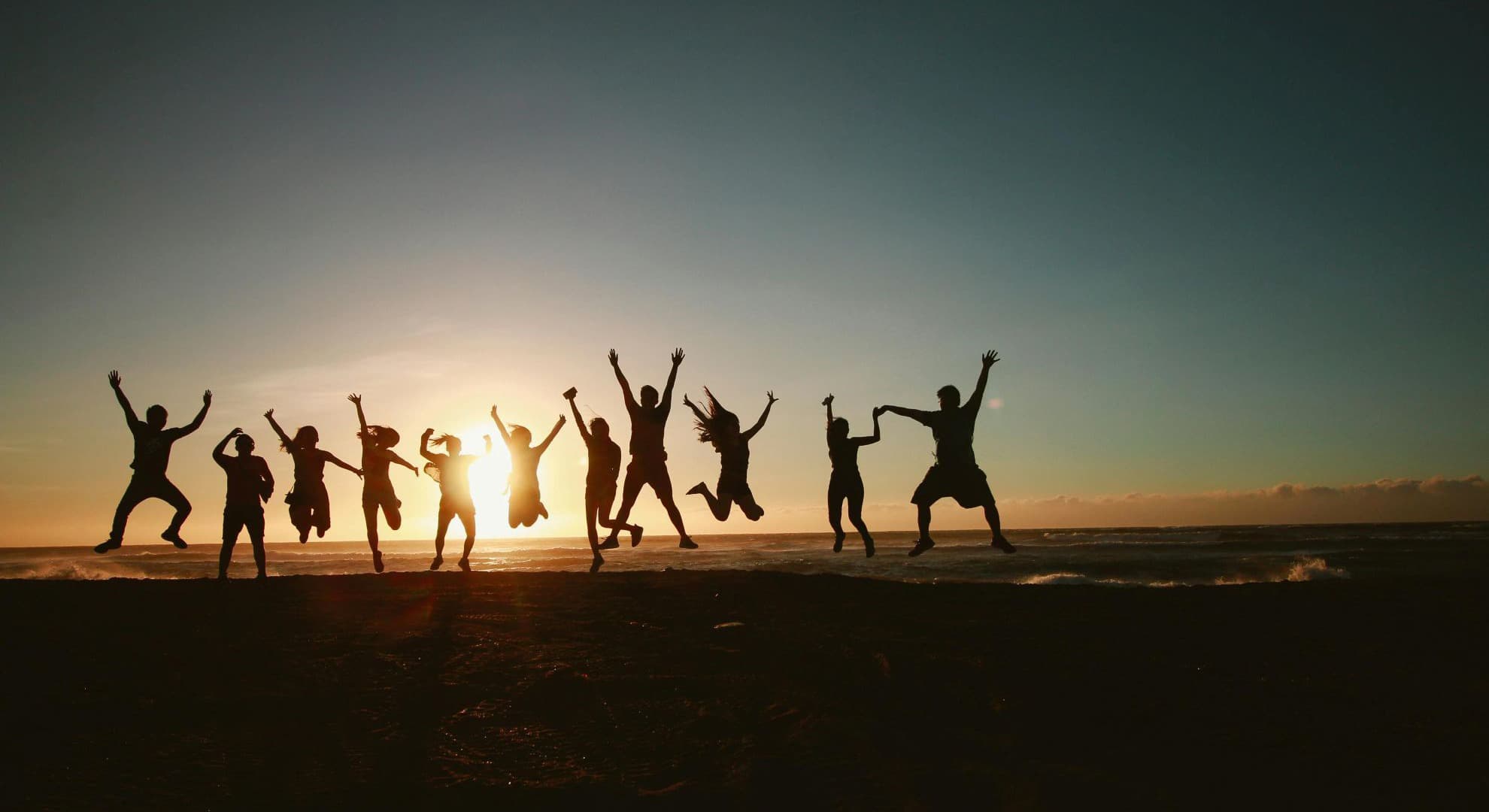 silhouette of 11 people jumping with hands in the air on a flat landscape against a sunset