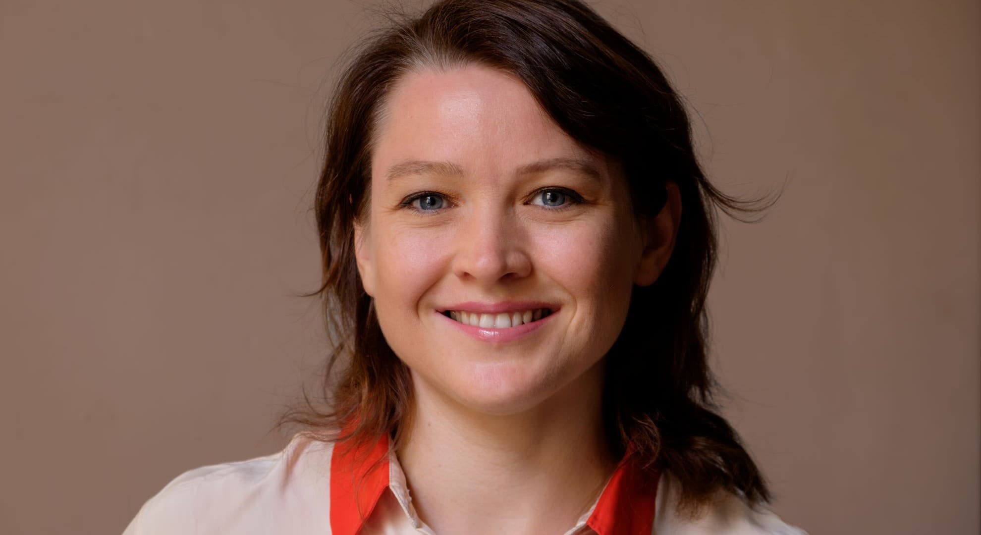 Close up head and shoulders person with brown shoulder length hair cream blouse with orange collar against light brown background