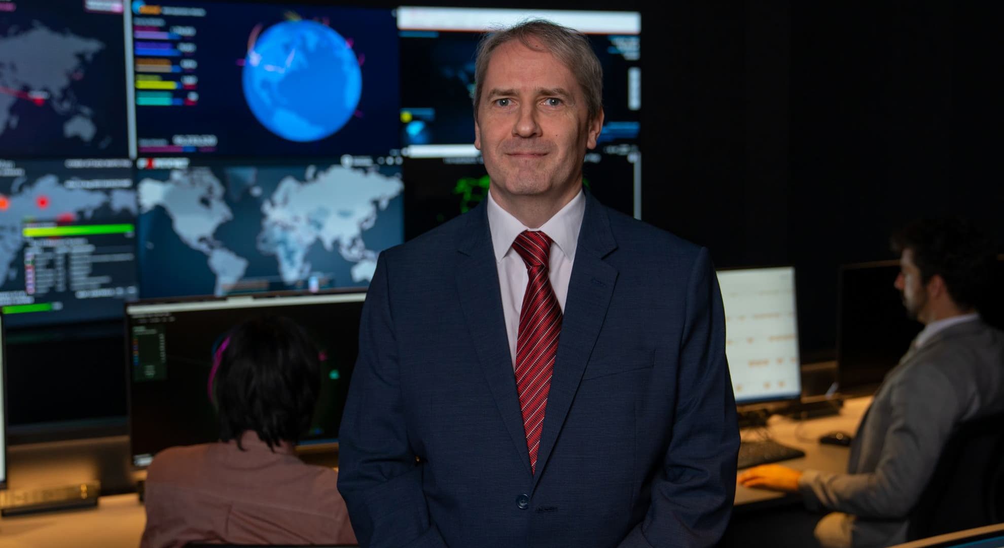 ECU's Professor Haskell-Dowland wears a suit and tie, behind him there are screens monitoring cyber security.