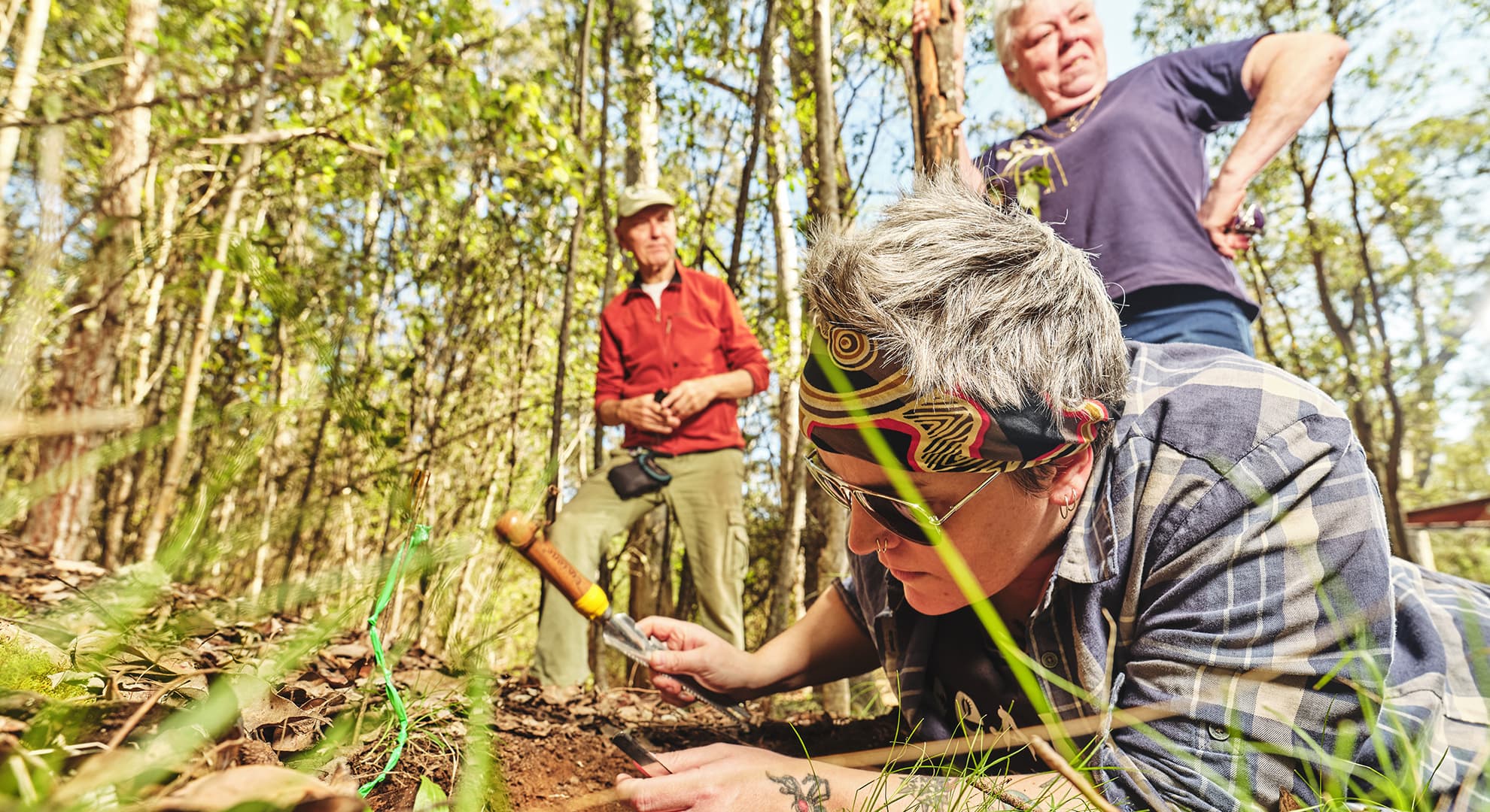 Person laying on the ground in the forest studying the earth with two people standing behind.