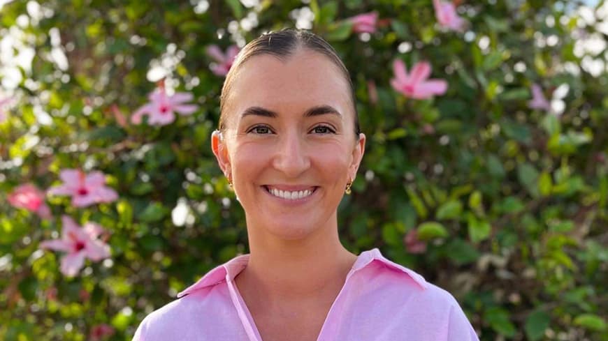 Nicolette Beard wears a pink button-down shirt, she grins outdoors in front of a green bush.