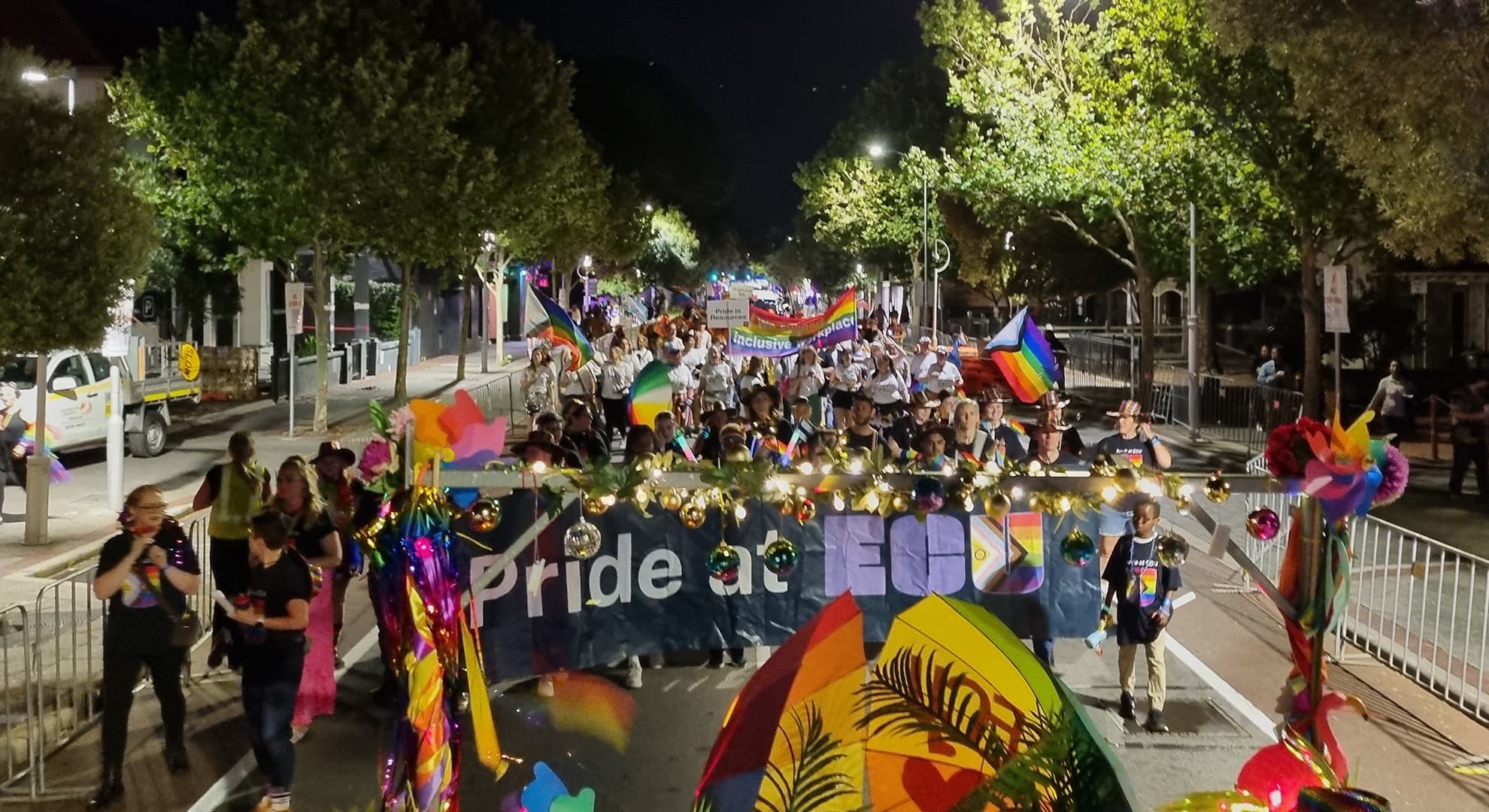 colourful crowd of people walking down a tree lined street carrying flags and a large banner that says Pride at ECU