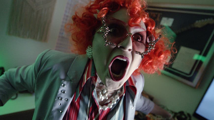 A person with red curly hair, white-painted face and pink sunglasses screams.