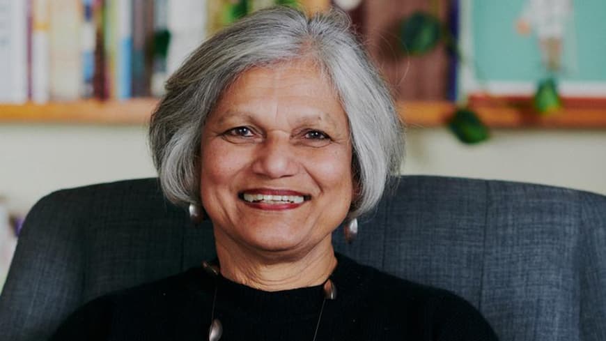 Caroline Wood AM wears a black sweater and sits on an armchair.