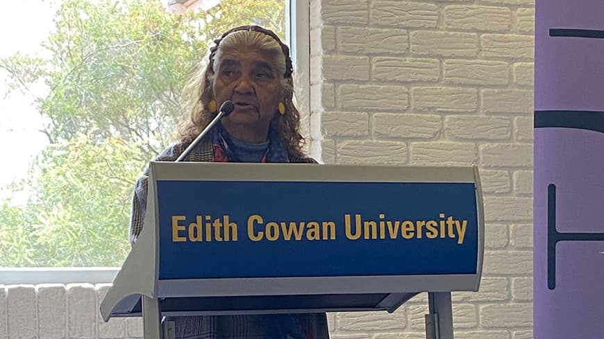 Elder Gloria Hill standing at Edith Cowan University lecturn against window with greenery outside and white wall