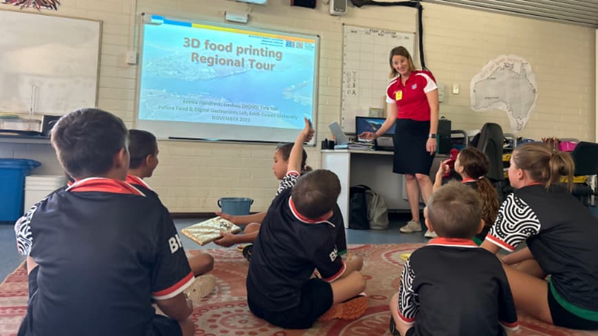 classroom setting with children in black uniforms with black and white Indigenous design on sleeves, sitting cross-legged on Indigenous design floor mat, watching a screen and listening to a presenter woman in a red polo shirt and black skirt.