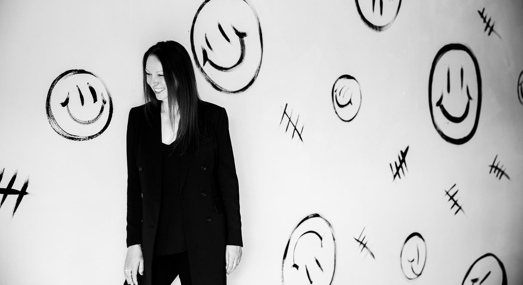 A black and white portrait of Professor Narelle Lemon, she is wearing a suit and smiles, there are smiley faces drawn on the wall behind her.