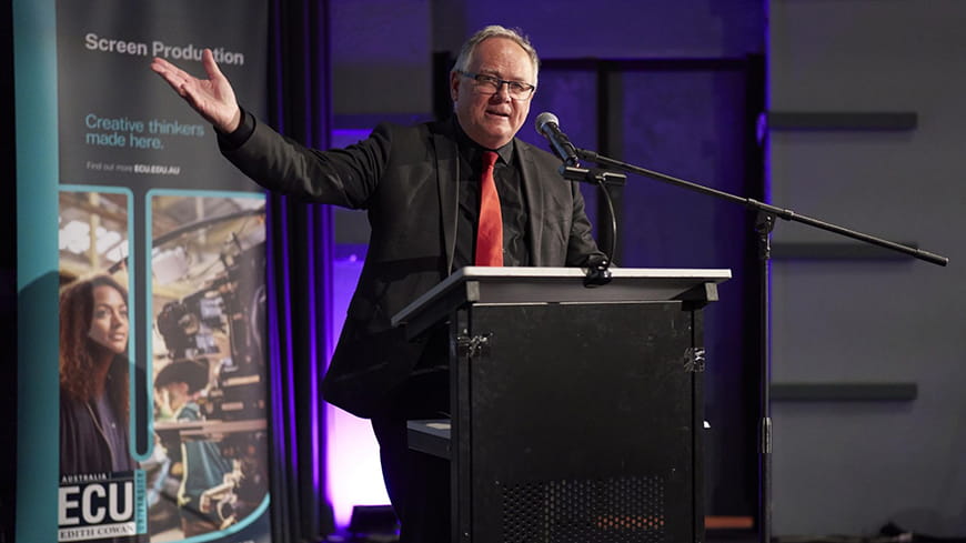 Man with grey hair and glasses speaking at podium in black suit and shirt with red tie, gesturing with his right arm in the air.