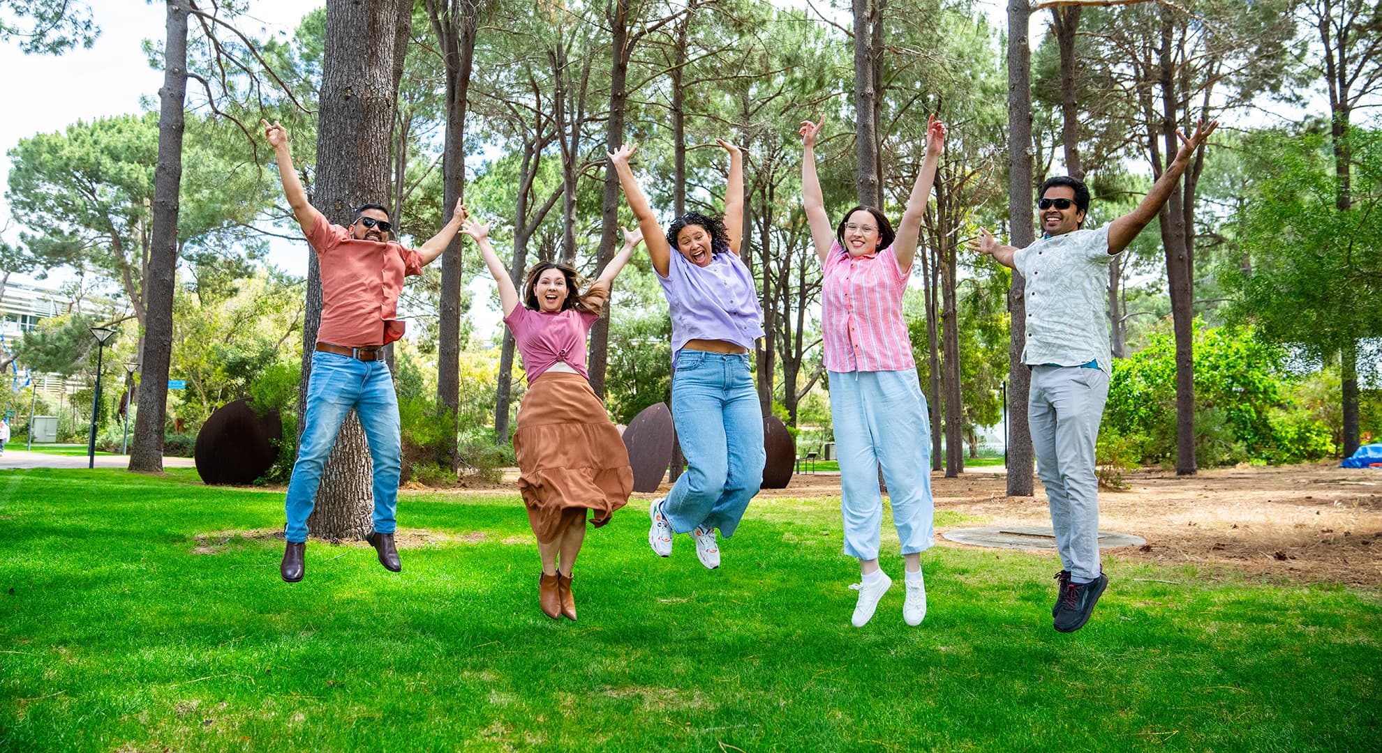 Five students jumping in the air on grass with pines trees in the background