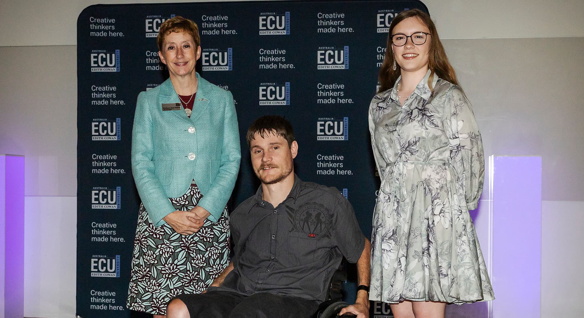 Three people in front of navy blue backdrop with ECU logos, person in centre in a wheelchair