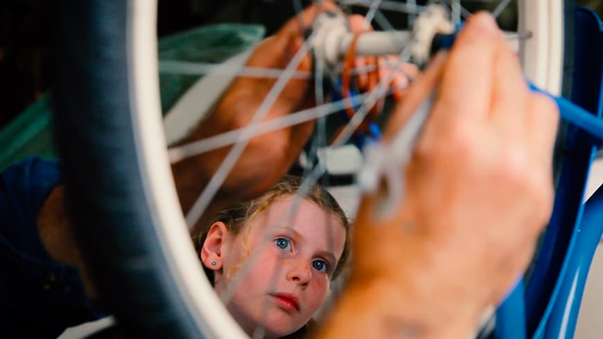 A young girl looks up at a bicycle wheel being fixed.