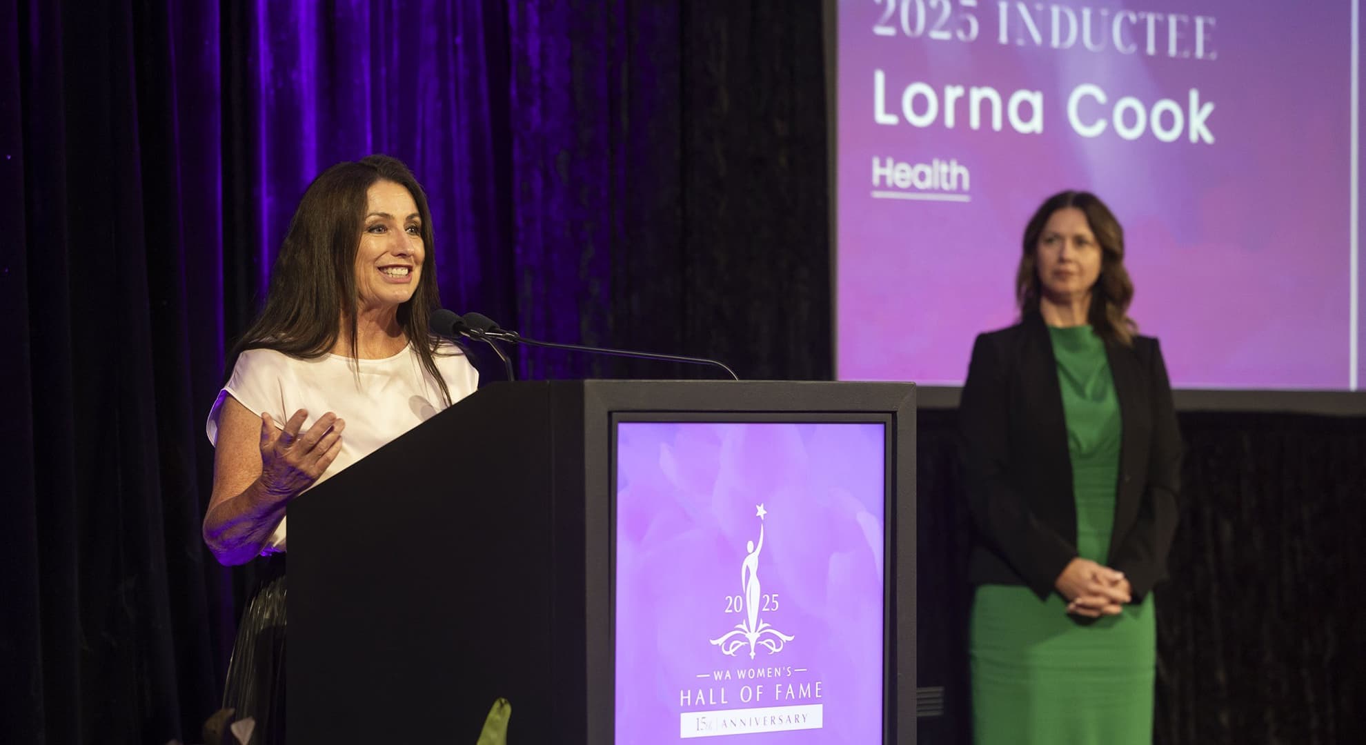 Person making a speech behind a lectern with a screen and another person stand on the right of the photo