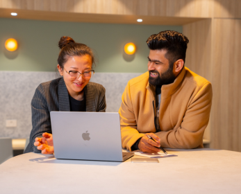 A woman in a dark grey jacket and a man in a tan coat sit and collaborate in front of a laptop computer in a modern workspace. 