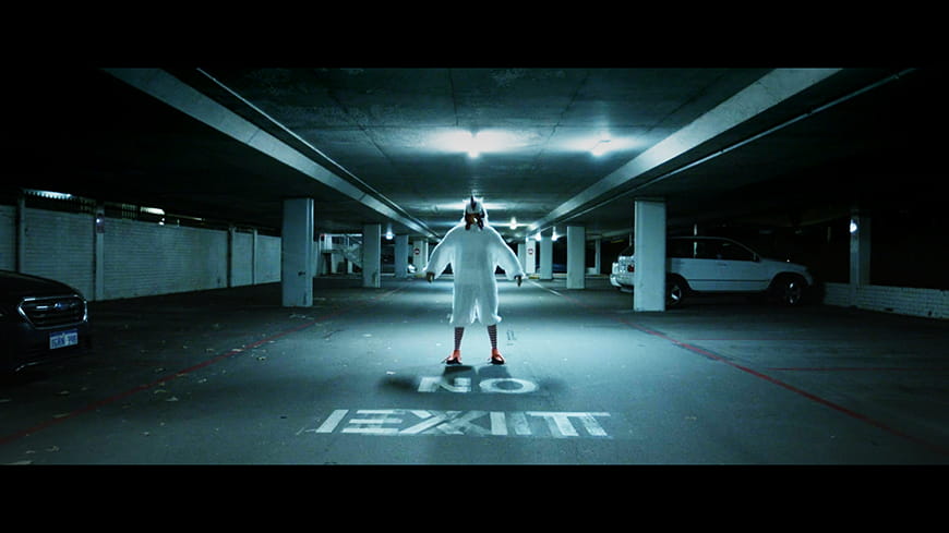 bleak, almost empty underground carpark, with person in a chicken suit standing staring at the camera in the centre with NO EXIT painted on ground.