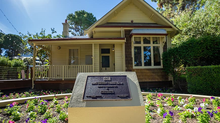 Traditional cottage house with flowering garden around plaque on a pedestal in middle of garden bed