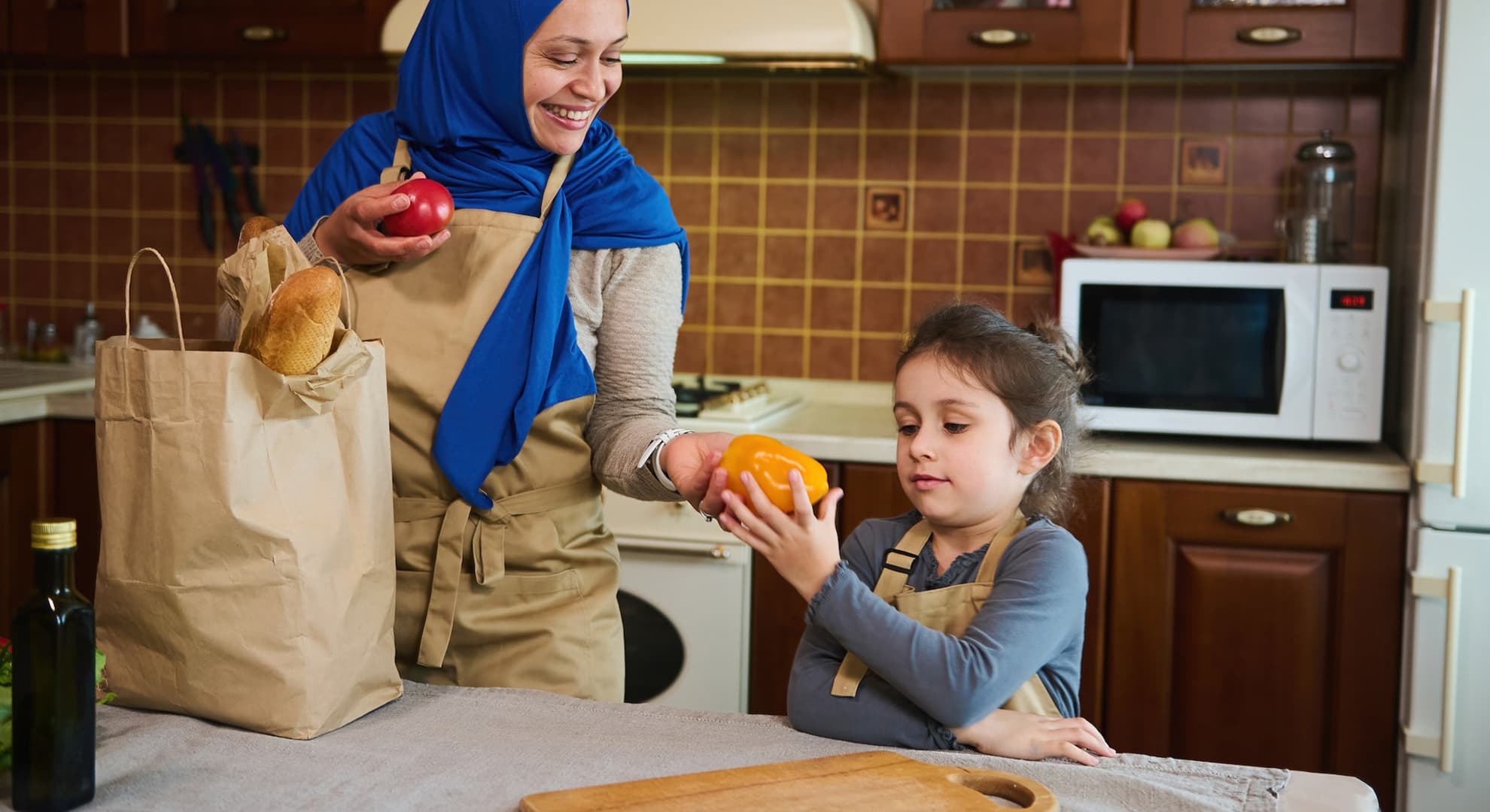 Woman in a kitchen wearing a hijab and smiling, handing a piece of food to a child.