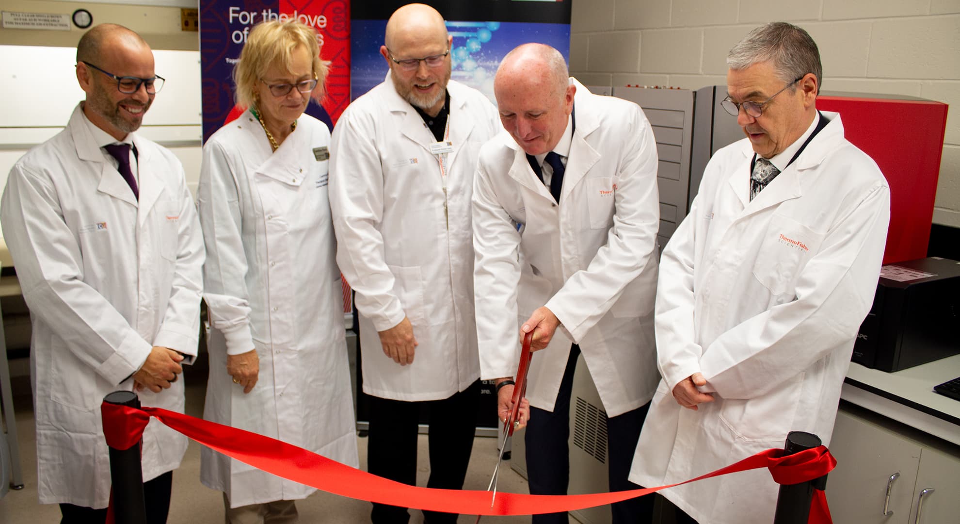 Five people in white lab coats cutting a red ribbon in front of laboratory equipment.