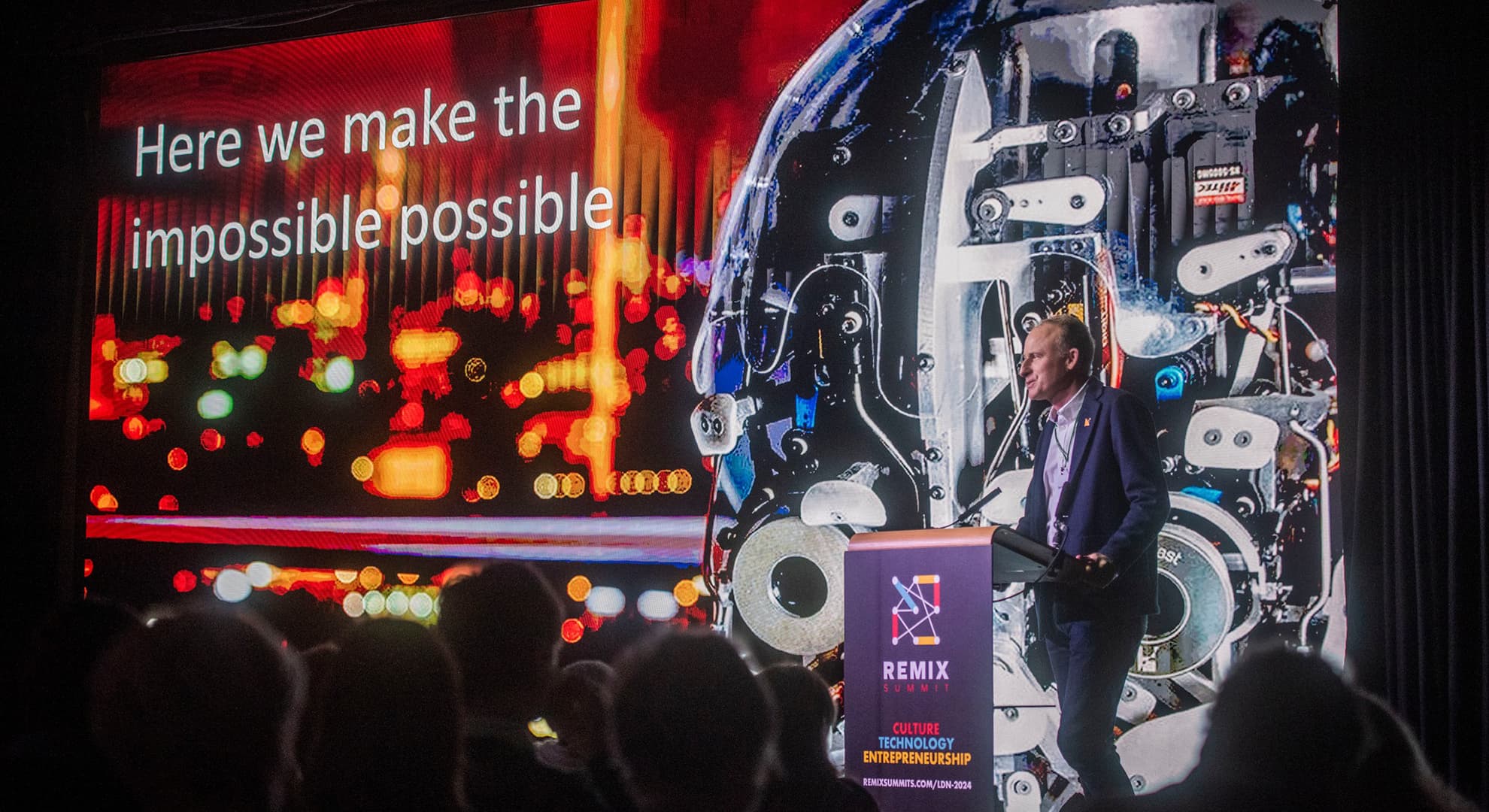 Large digital screen with electronic imagery with person standing in front at a lectern branded REMIX