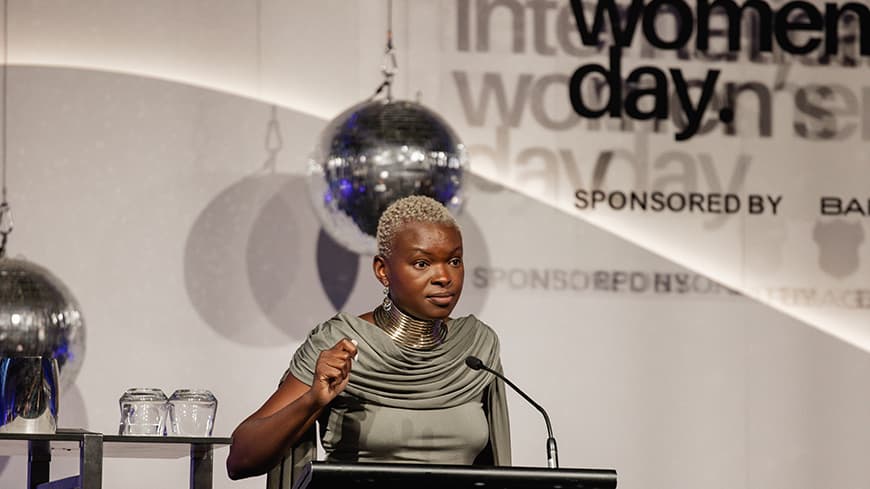 Person speaking at lectern on stage in front of International Women's Day backdrop.